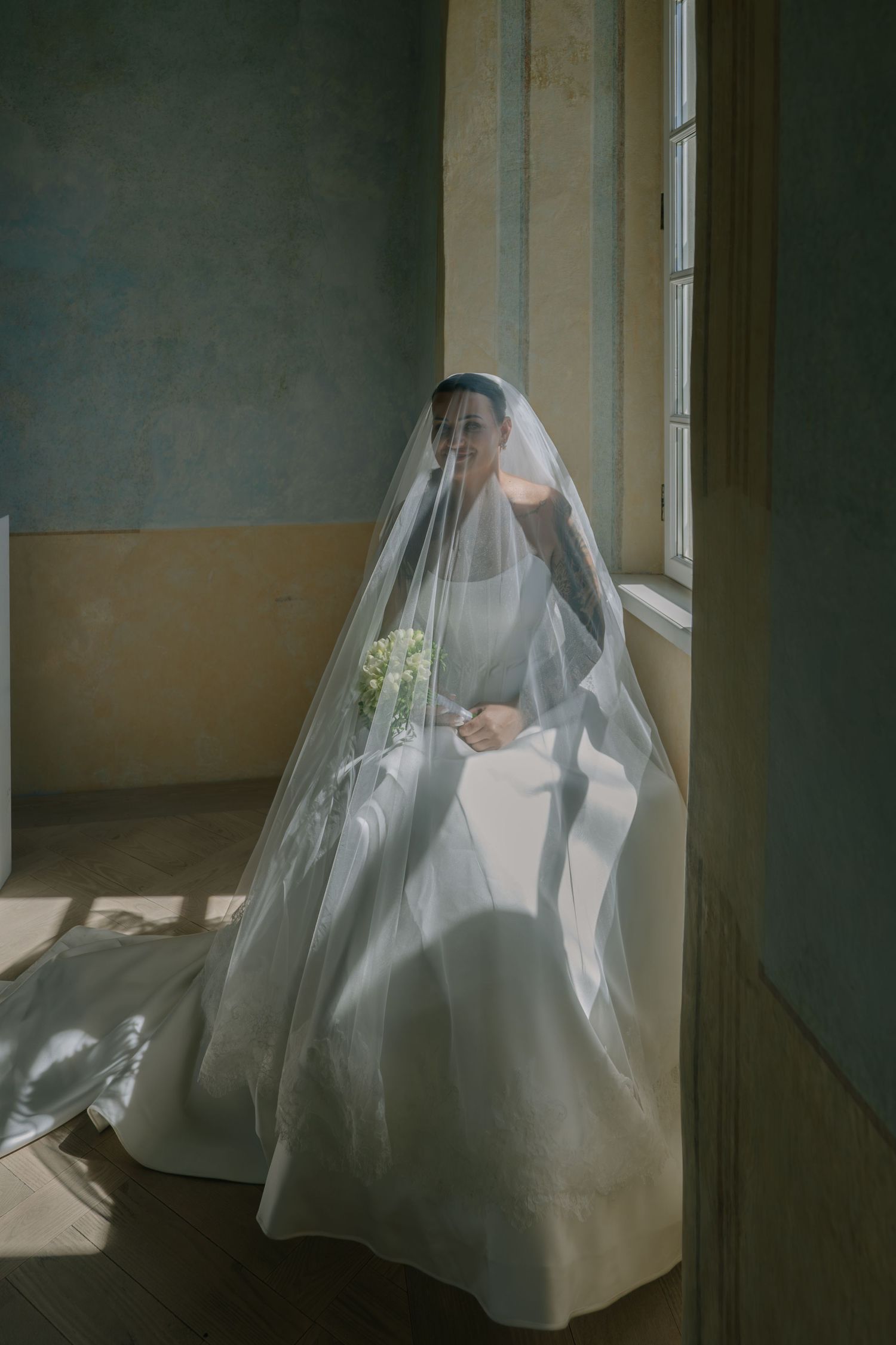 Bride in a flowing white gown and sheer veil sits holding a bouquet in a sunlit room with softly weathered walls.