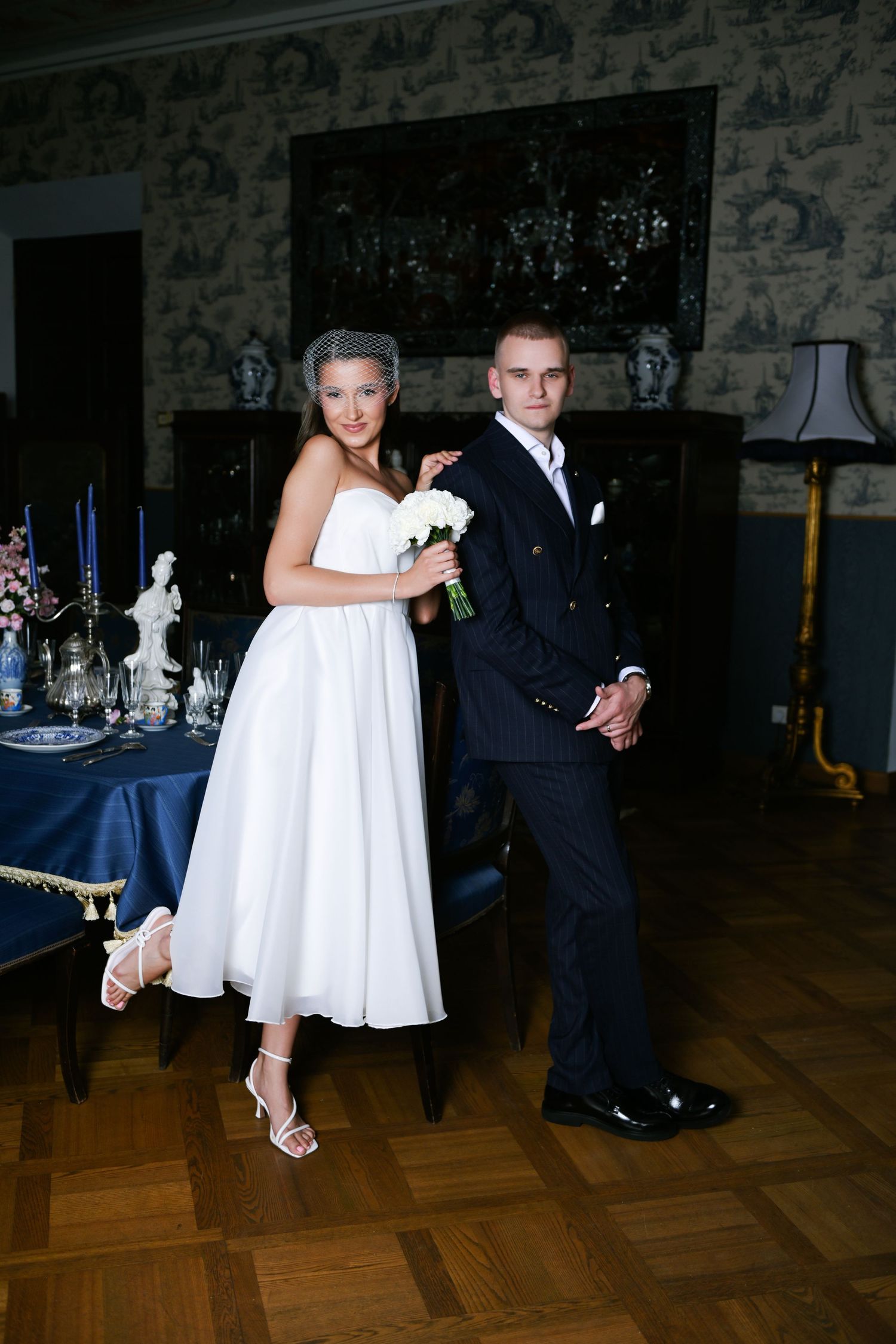 Bride in a white tea-length dress holding a white bouquet poses beside her groom in a navy pinstripe suit in an elegant, vintage-style room.