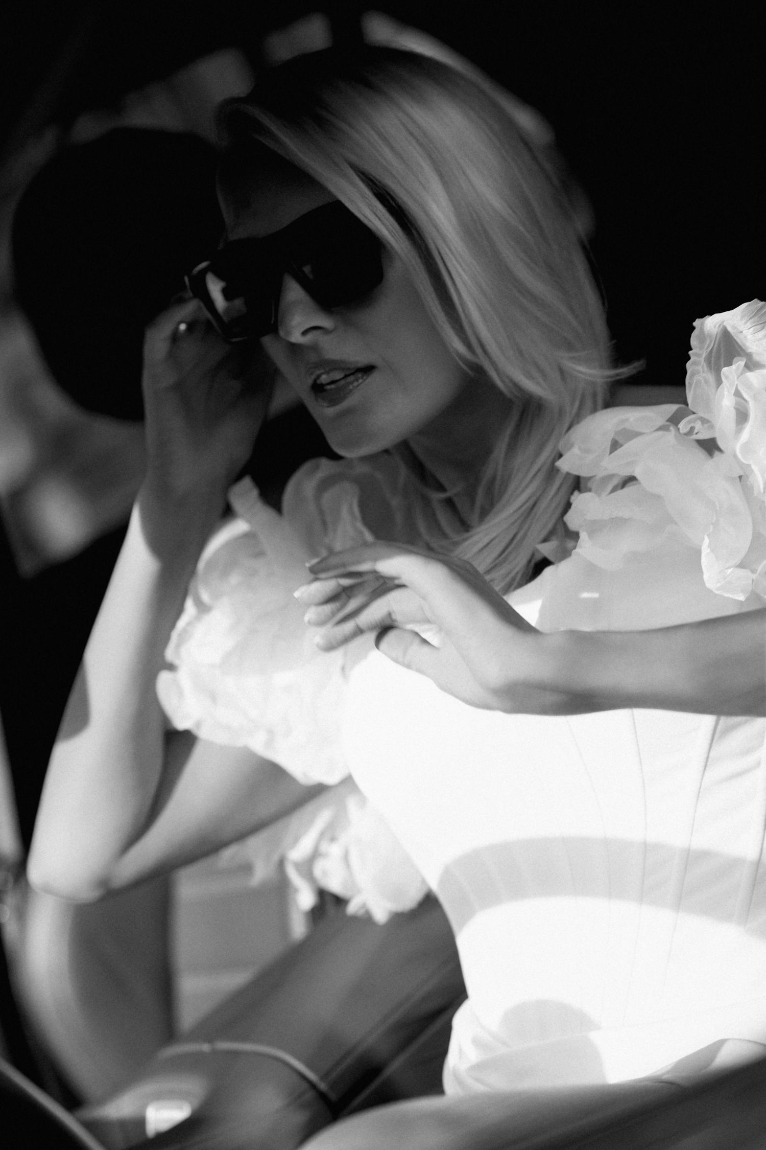 Black and white photo of a blonde woman in a white ruffled off-the-shoulder top adjusting her dark sunglasses while seated in a car.