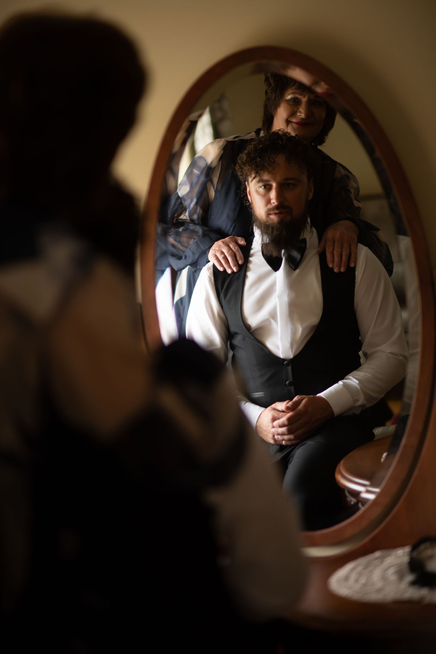 Groom in vest and bow tie sits before oval mirror as bride adjusts his collar in warm romantic light.