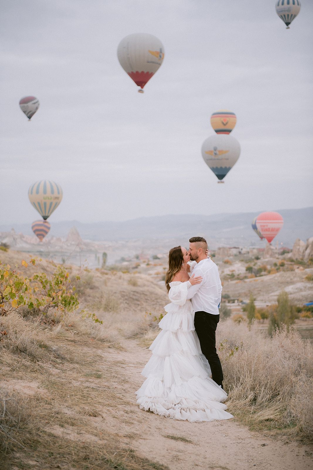 Hot air balloons float in the sky over Cappadocia as a couple shares a romantic moment on a hillside path.