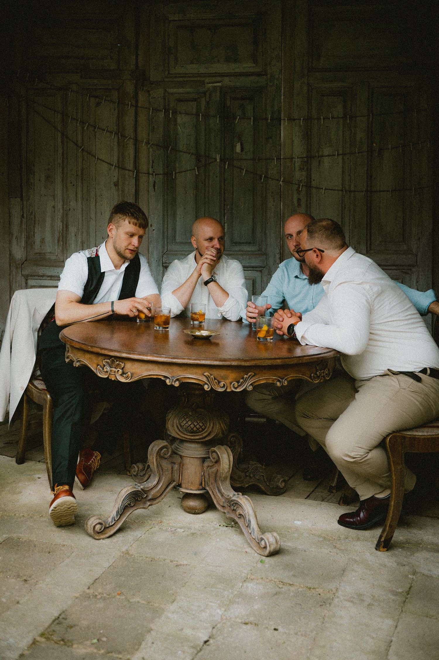 Two men in white shirts engage in an arm wrestling match at an ornate wooden table in a rustic setting.
