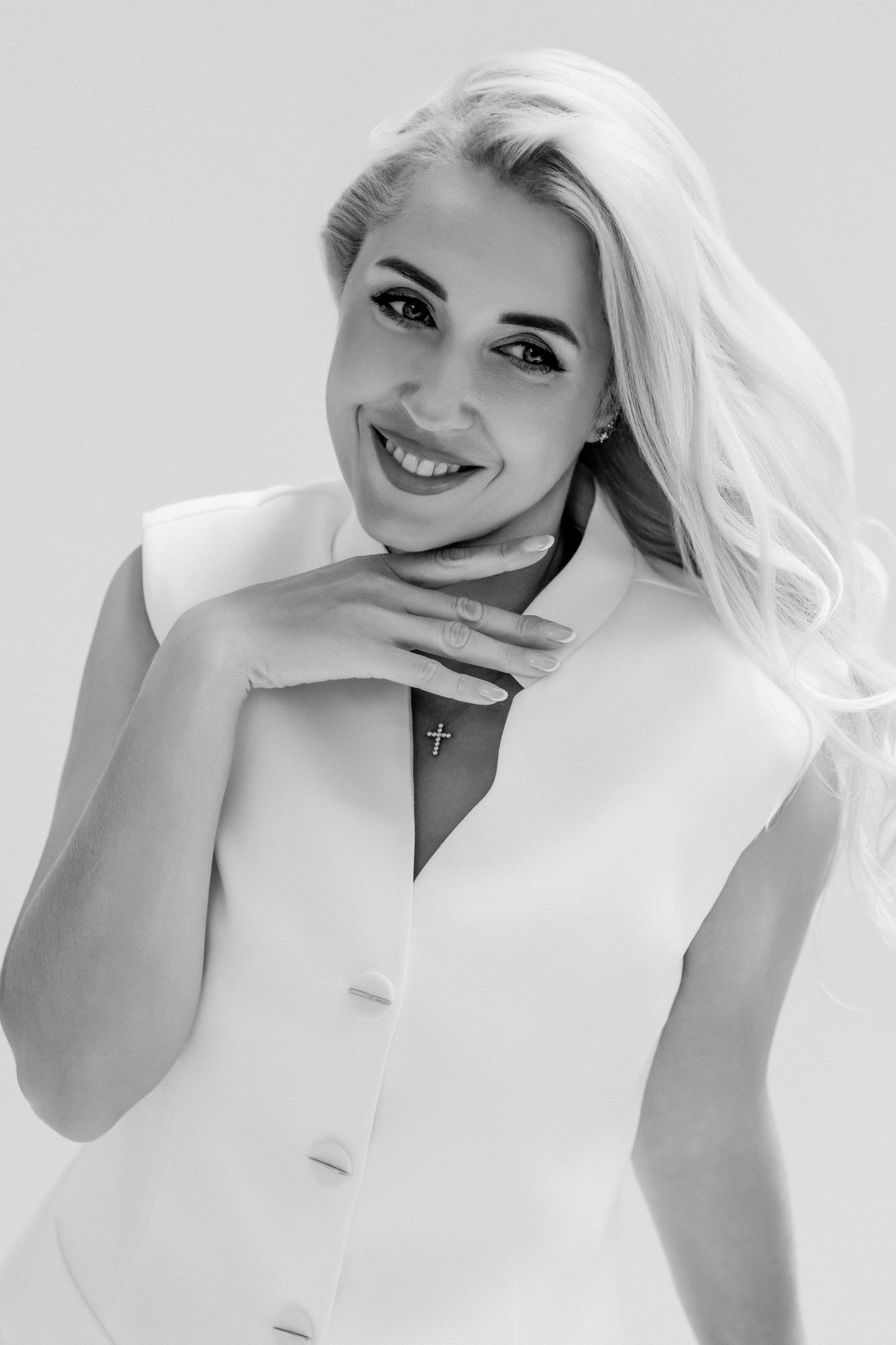 Smiling blonde woman in white sleeveless blazer wearing cross necklace, black and white portrait photo.