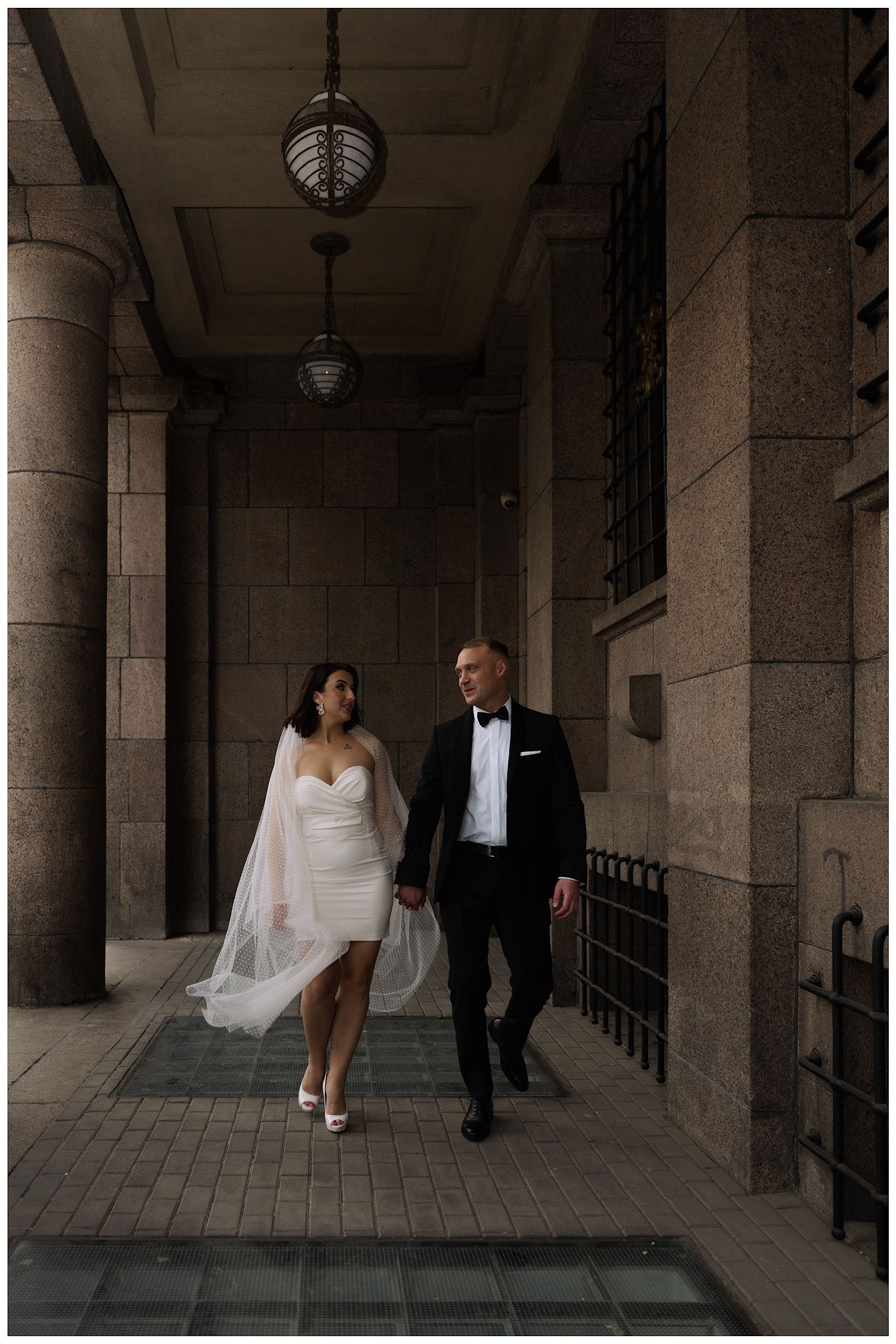 A bride and groom stroll through a grand columned hallway with classic architectural details.