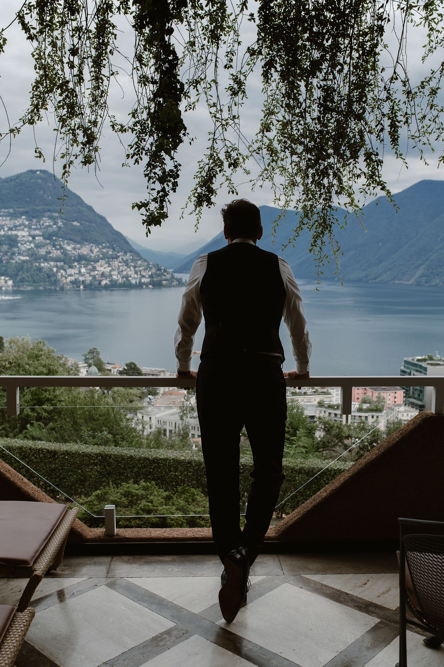 A silhouetted figure stands on a terrace overlooking a scenic mountain lake view with lush greenery and distant hills.