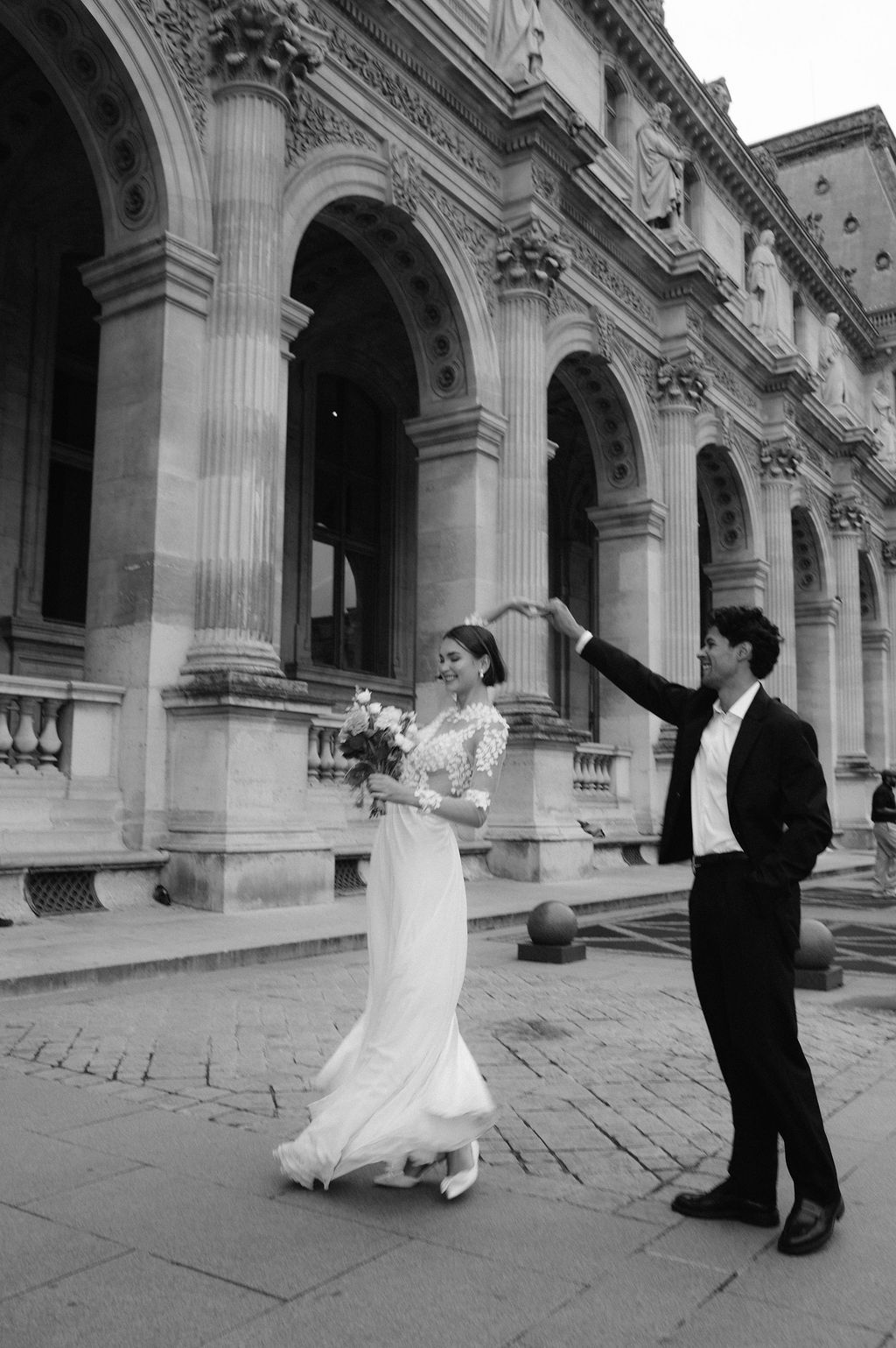 Black and white photo of a bride and groom dancing outside a grand classical building, bride holding a bouquet.