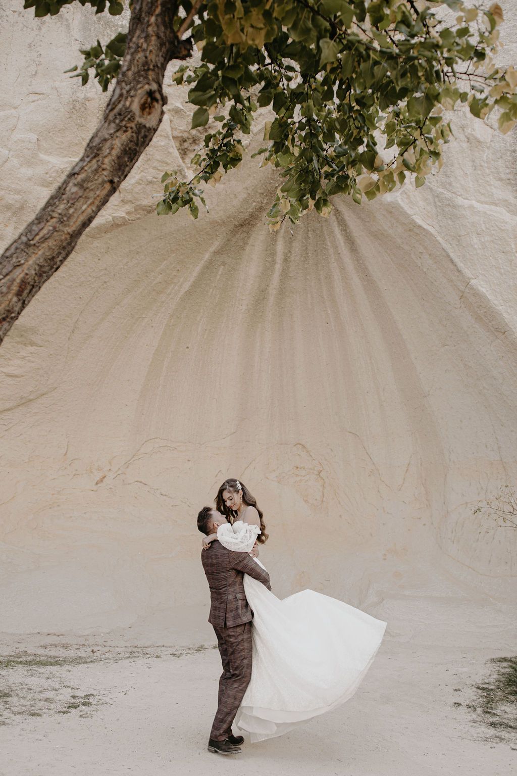 Groom lifts bride in white flowing dress near sandy rock formation and tree in romantic outdoor wedding photo.