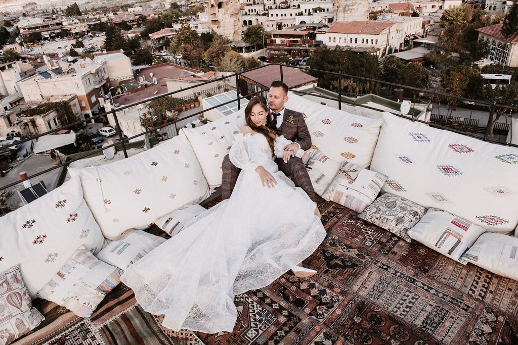 Couple in wedding attire posing on a rooftop with city views, bride in white gown, groom in brown suit.