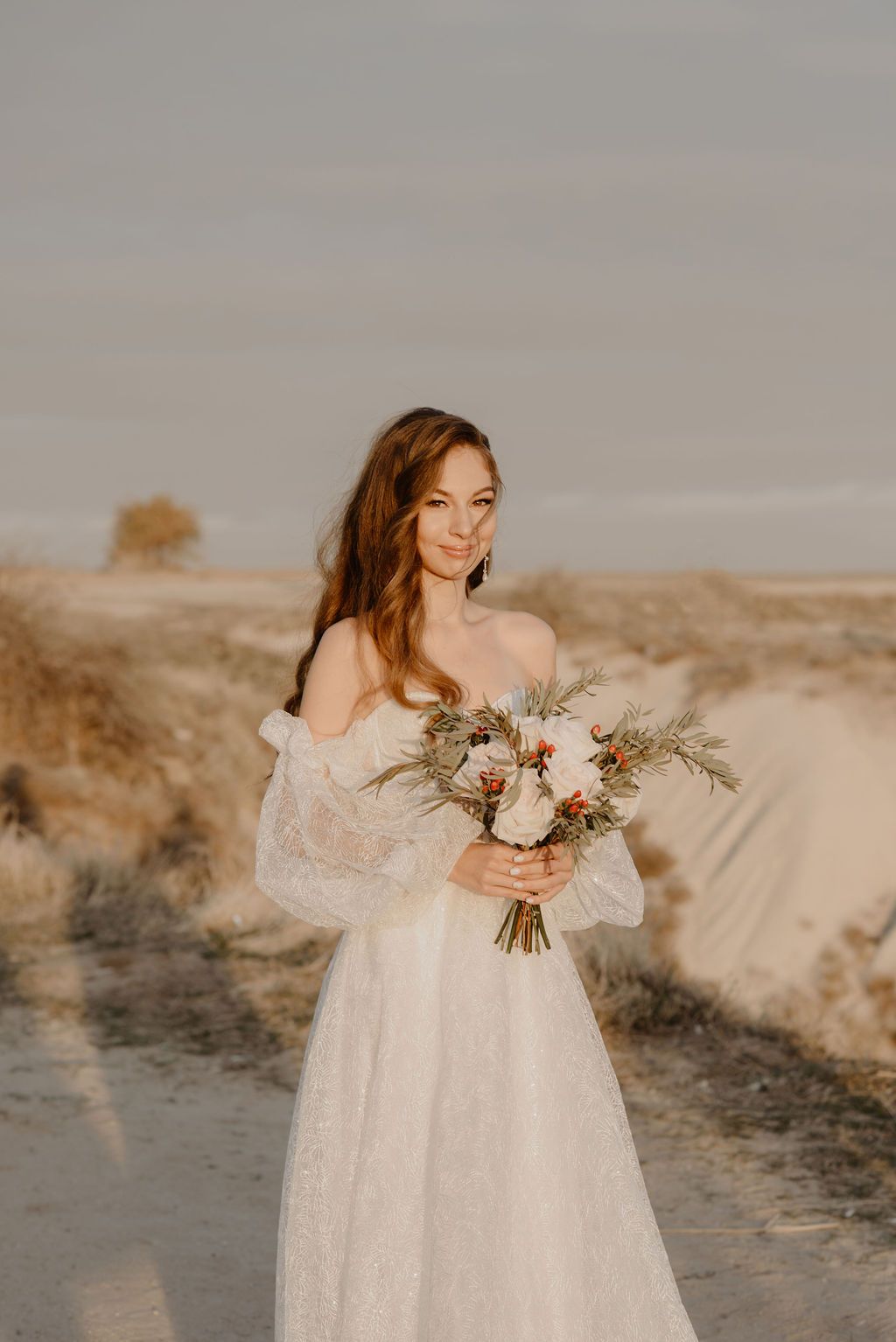 Bride in off-shoulder lace gown holds boho bouquet in sandy desert landscape at golden hour.