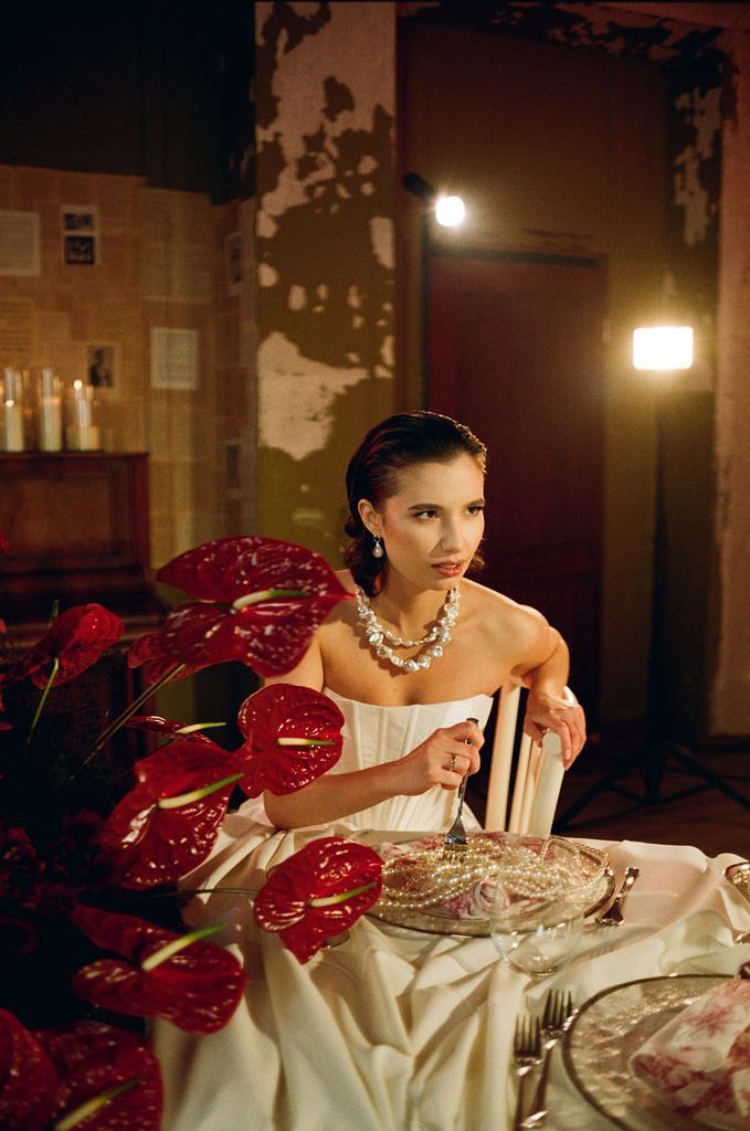 Elegant woman in white strapless gown with pearl necklace seated at a candlelit table with red anthurium flowers.