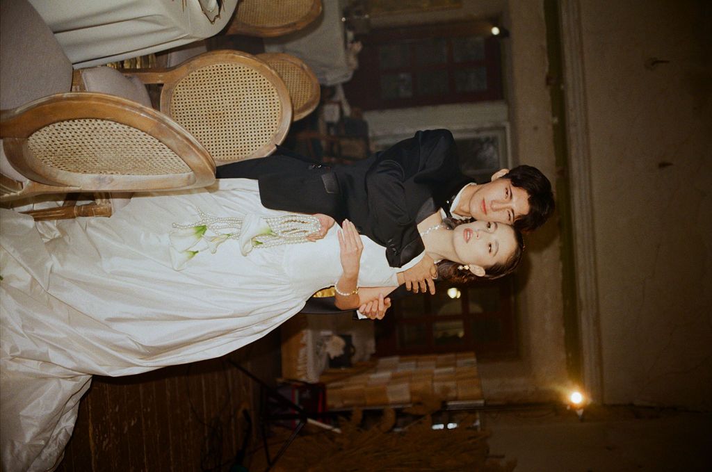 Couple embracing romantically outdoors, bride in white floral dress and groom in black outfit at a rustic setting.