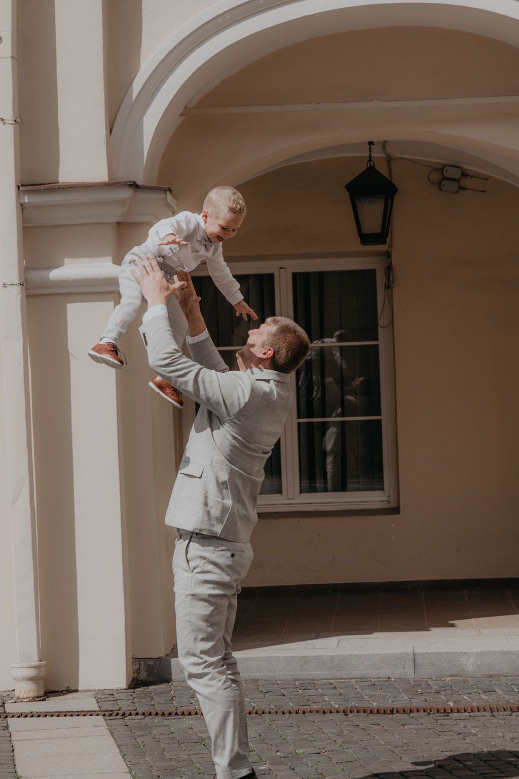 Father in gray suit tossing smiling baby in air outside classic European building.