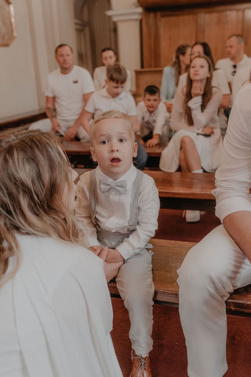 Adorable young boy in white suspenders and bow tie sits on church pew during a family ceremony.