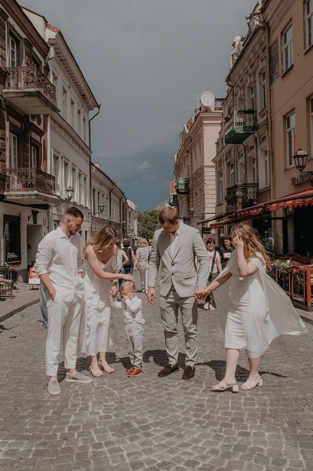Family in white outfits holding hands on a cobblestone street in a European city on a sunny day.