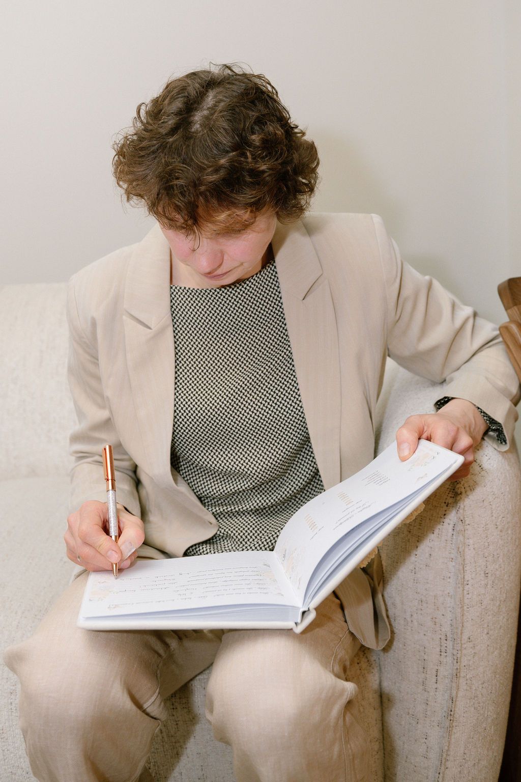 Curly-haired man in beige blazer writing in open notebook while seated on sofa.