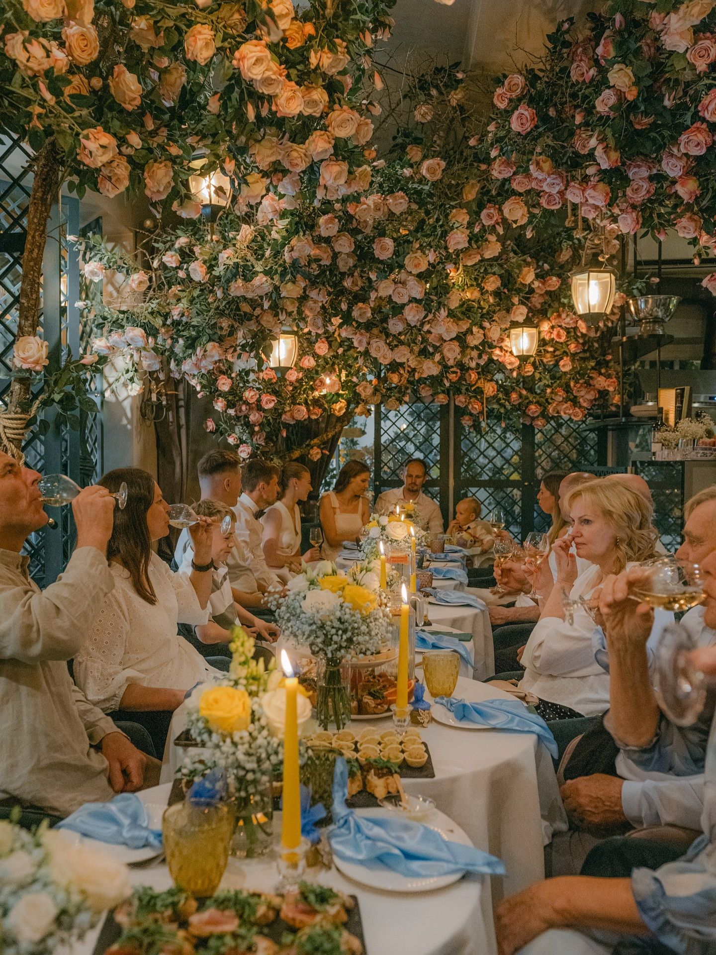 Elegant wedding reception with floral ceiling installation of pink roses, candlelit tables, and guests in white attire.