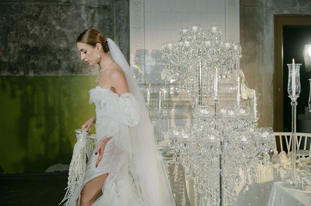 Elegant bride in white off-shoulder gown and veil stands beside crystal candelabra centerpiece at luxurious wedding reception.