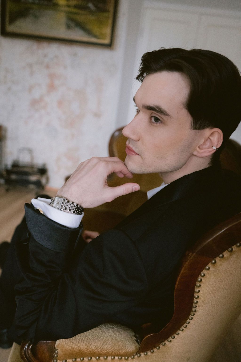 Young man in black suit sitting thoughtfully in armchair, wearing a watch, with elegant interior background.