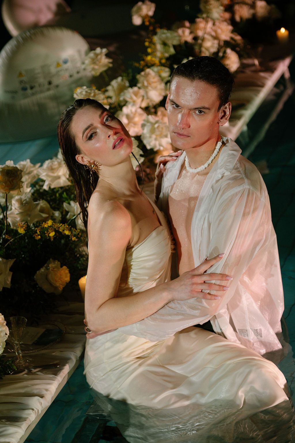 Elegant couple in white attire posing romantically among yellow flowers in a dreamy, candlelit setting.