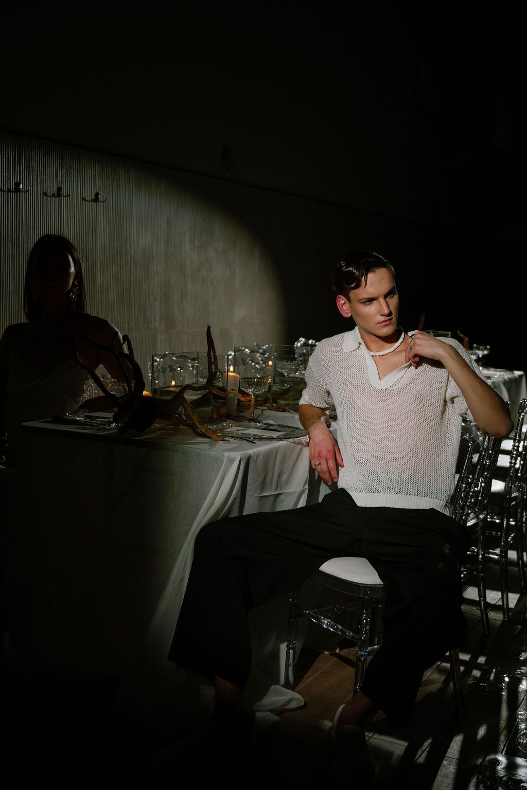 Young man in white shirt sits at candlelit dinner table in dramatic dark setting with another guest nearby.