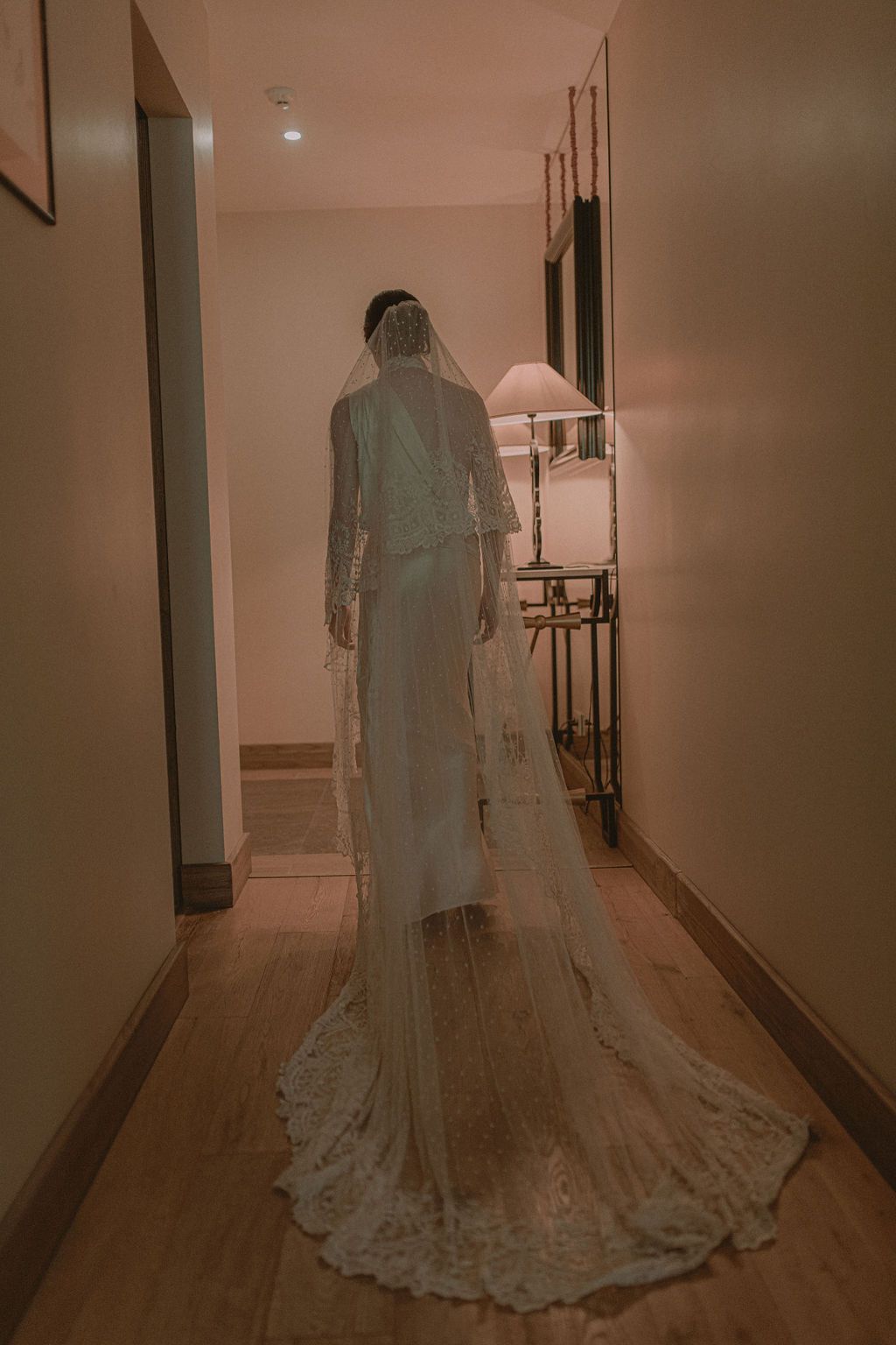 Bride in elegant lace wedding gown with long veil stands in warmly lit hotel corridor.