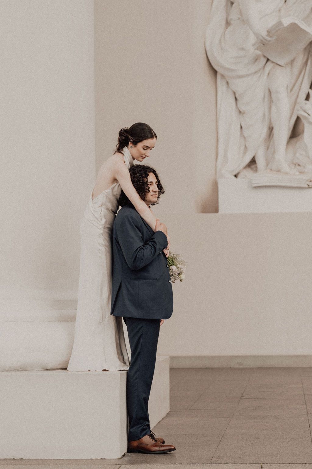 Bride in white gown embraces groom in navy suit near marble statue in elegant wedding portrait.