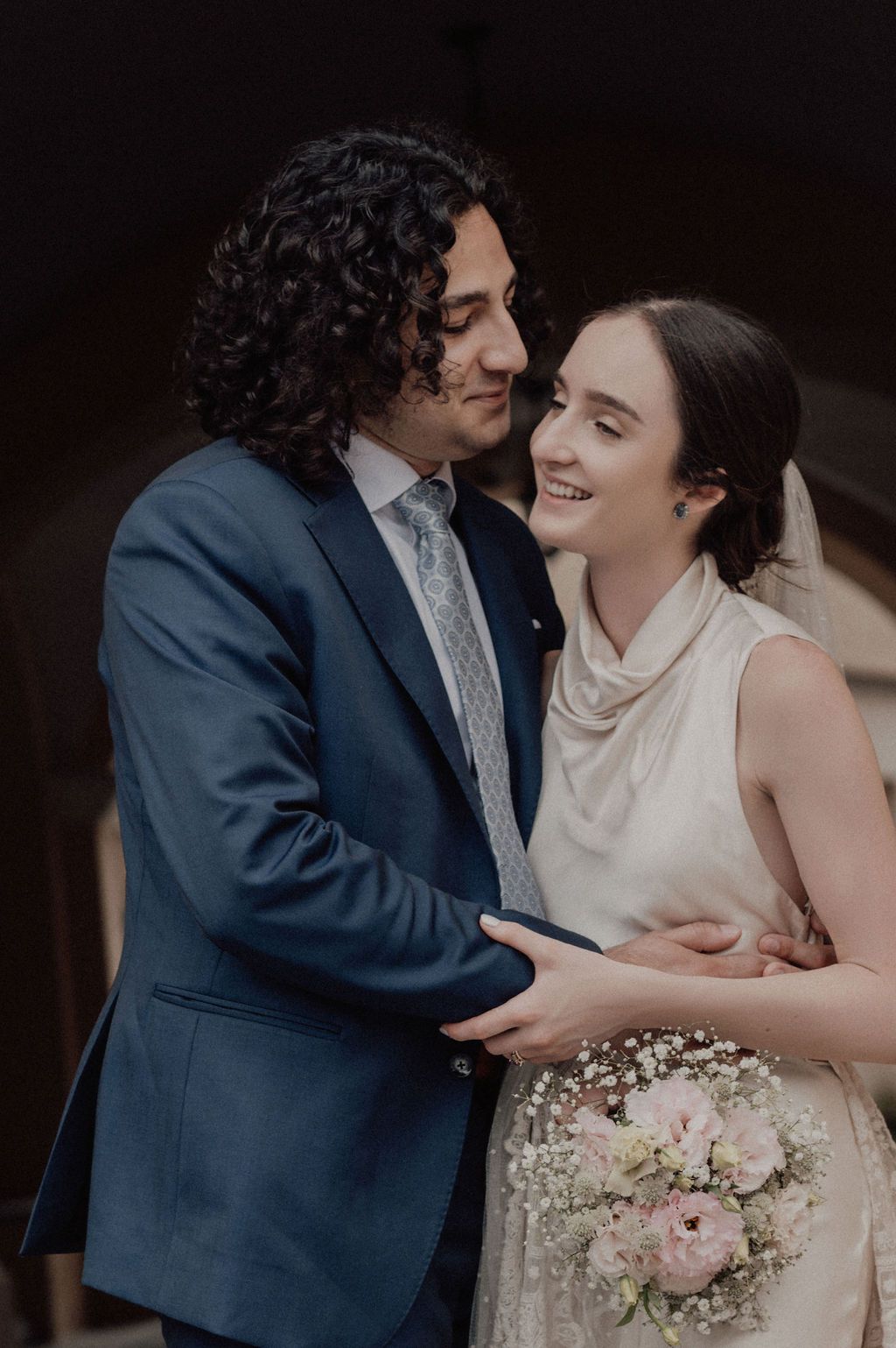 A smiling bride holding a pink bouquet embraces a groom in a blue suit during their wedding day.