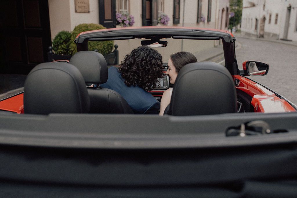 Couple sharing a romantic moment in a red convertible car on a cobblestone street.