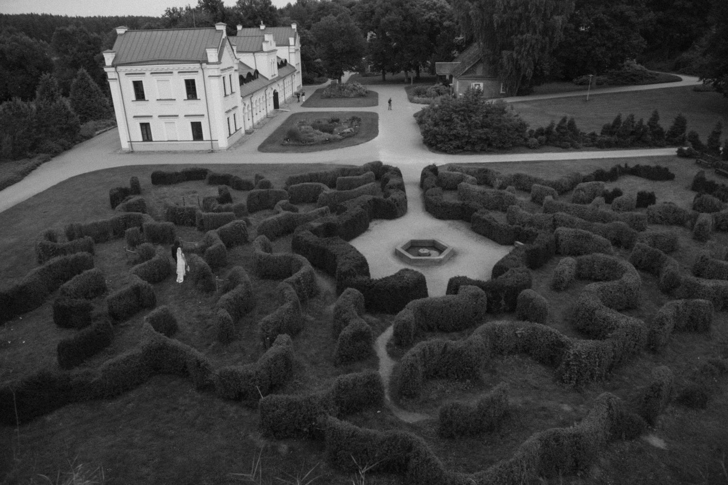 Black and white aerial view of a historic manor house overlooking a formal garden with sculpted hedge mazes and a central fountain.