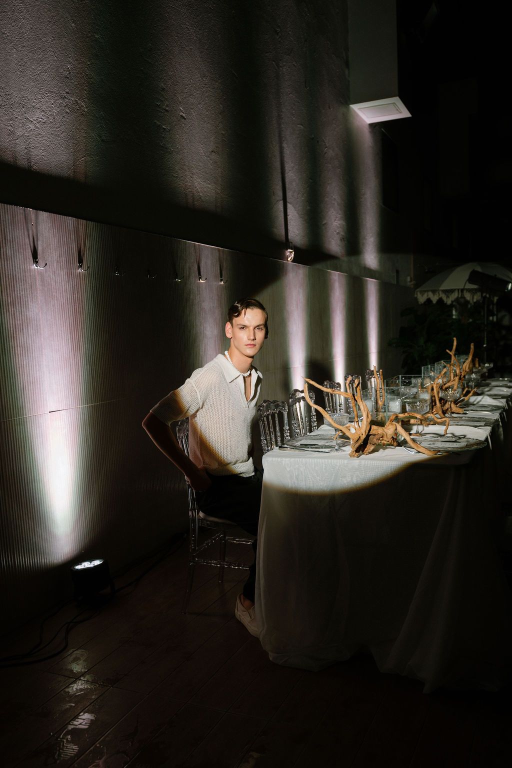 Man in a light button-up shirt sits beside a long banquet table with driftwood centerpieces in a dimly lit room with dramatic wall lighting.