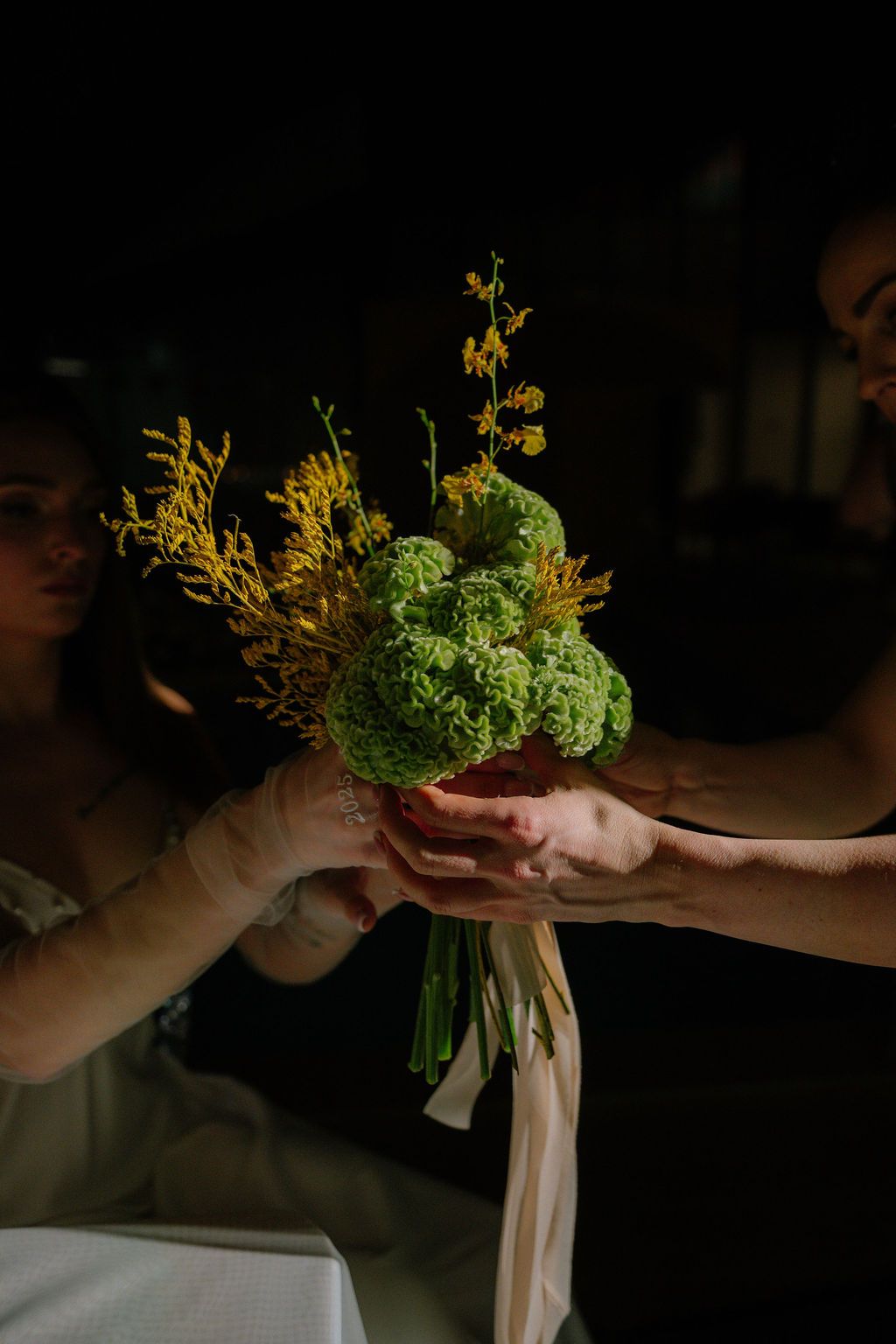 Two hands exchange a bridal bouquet of green viburnum and yellow wildflowers tied with a cream ribbon against a dark, moody background.
