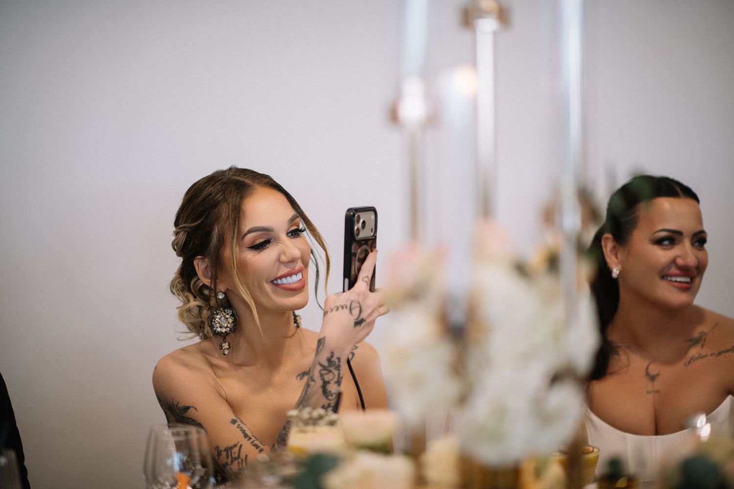 Smiling woman with an updo and arm tattoos takes a photo with her phone at a wedding reception table beside another woman.