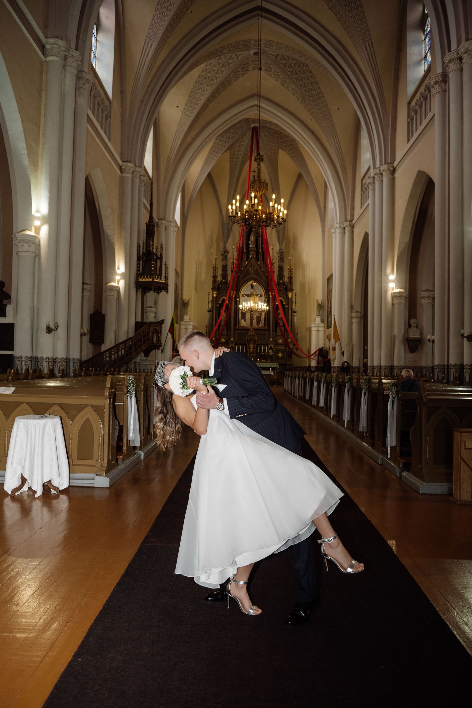 Bride in a white tea-length dress and groom in a dark suit share a dramatic dipped kiss on the aisle of an ornate Gothic church.
