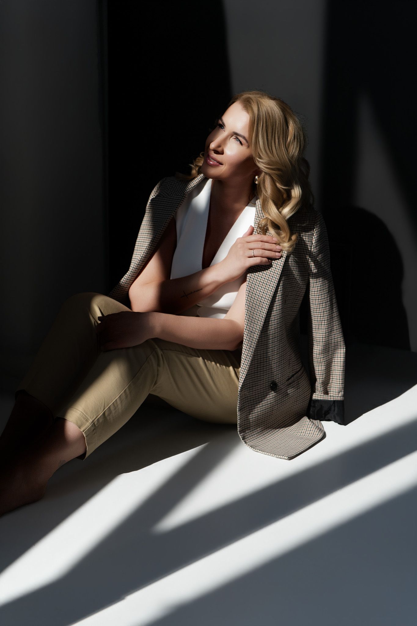 Blonde woman in beige trousers and blazer sitting on floor in dramatic sunlight and shadows.