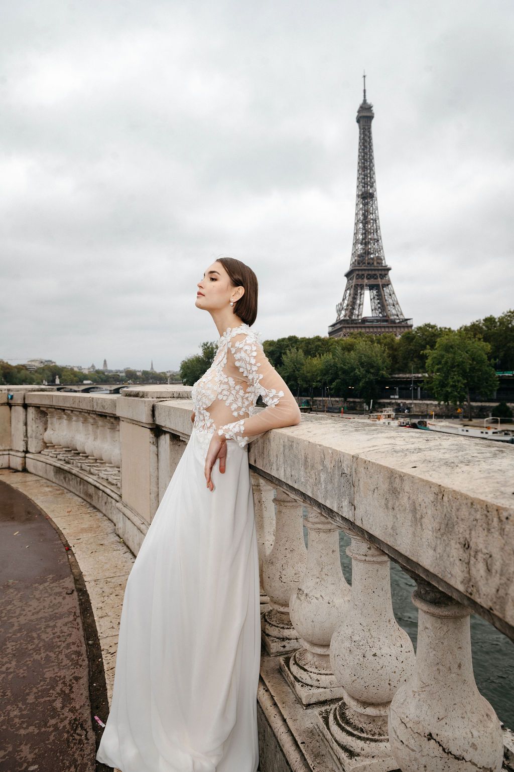 Bride in white lace gown poses on stone bridge balustrade with Eiffel Tower in Paris background.