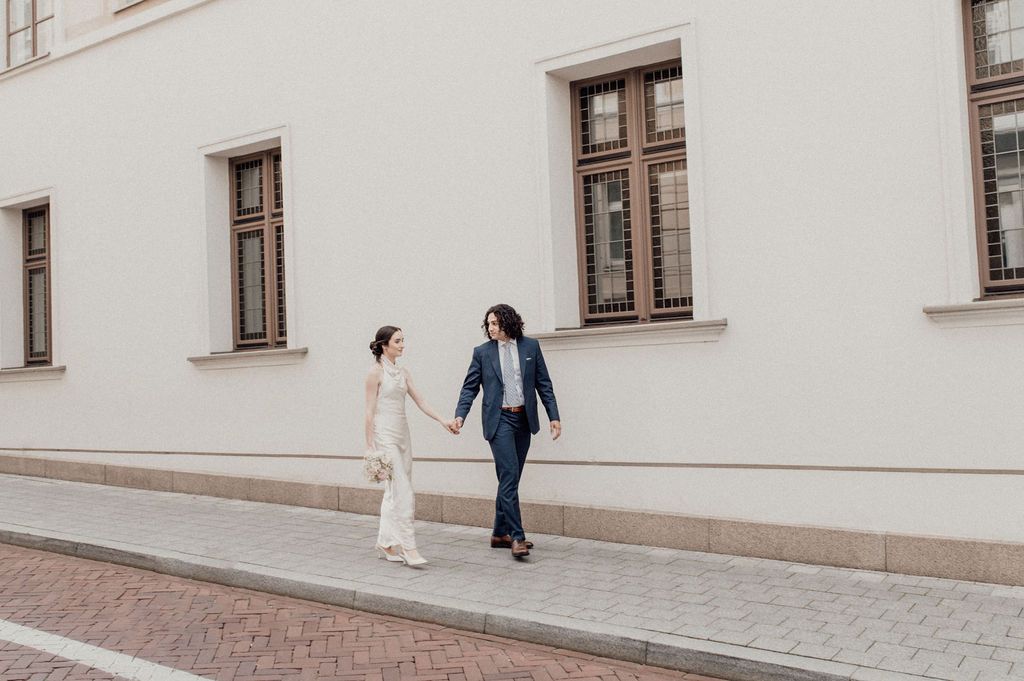 Couple holding hands walking along a cobblestone street beside a white building with brown windows.