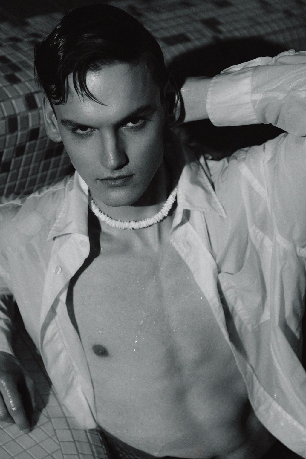 Black and white photo of a young man in wet open white shirt with pearl necklace posing by a pool.