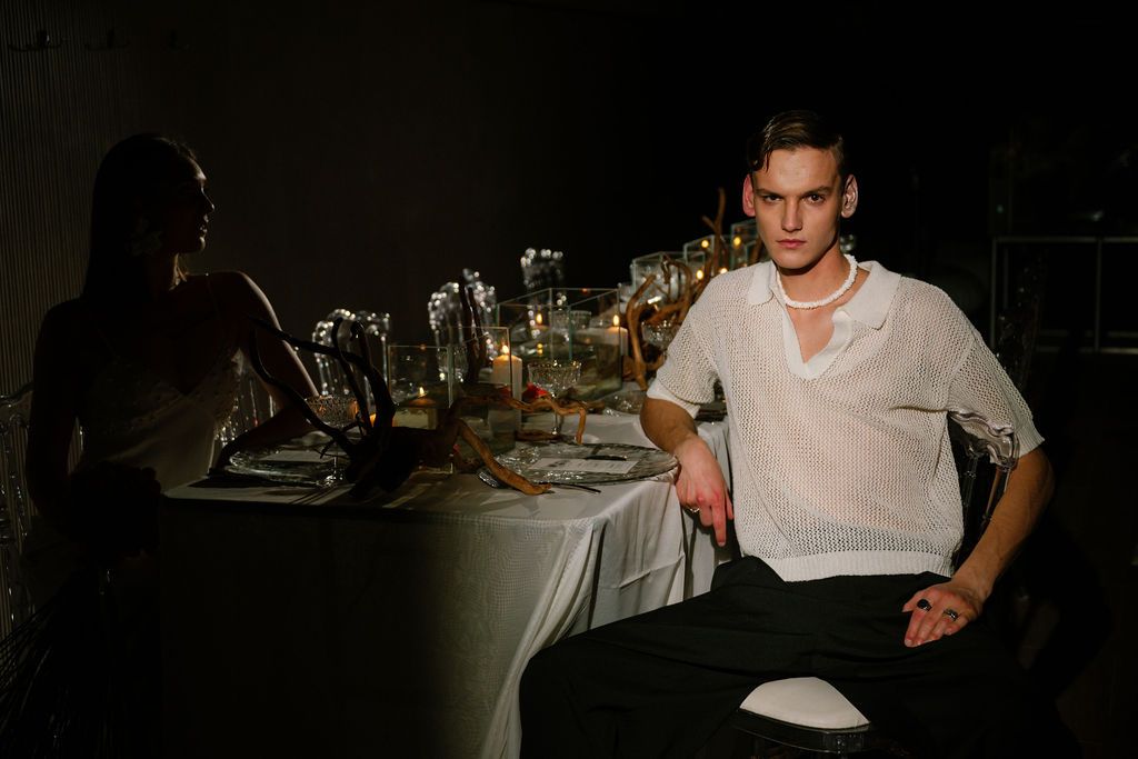 Young man in white sheer shirt and pearl necklace seated at an elegantly set dinner table in dim lighting.