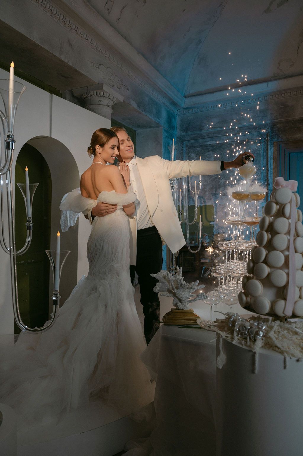Bride and groom cutting a sparkling wedding cake in an elegant venue, bride in mermaid gown with off-shoulder wrap.