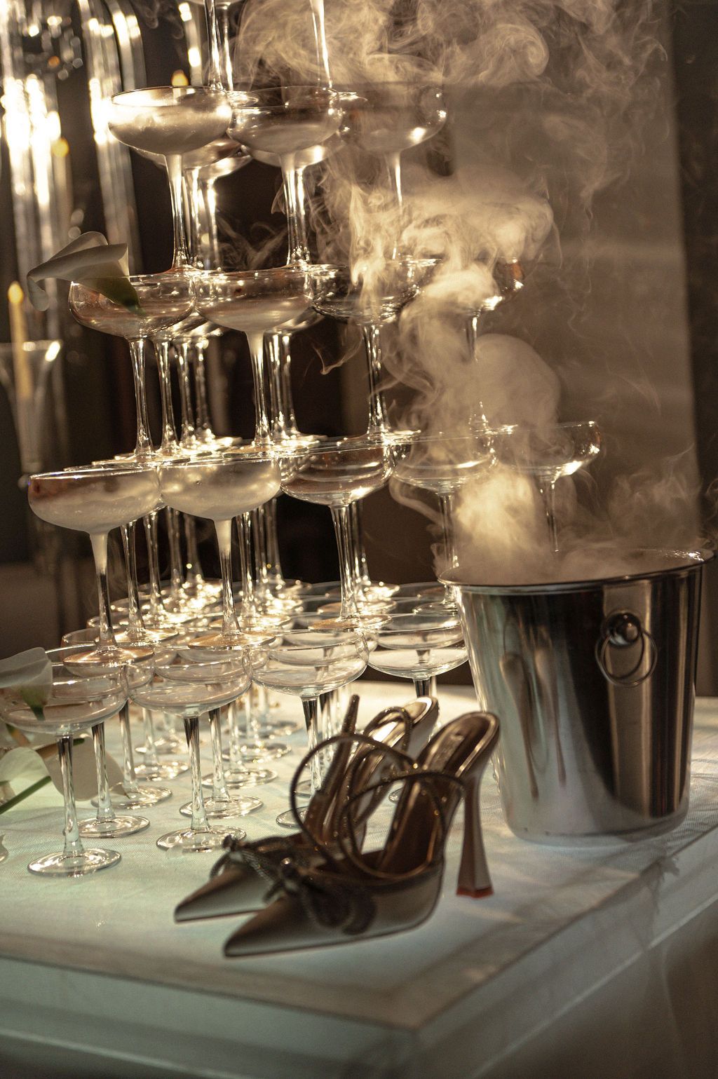 A champagne tower of coupe glasses beside strappy heels and a smoking ice bucket at an elegant party.