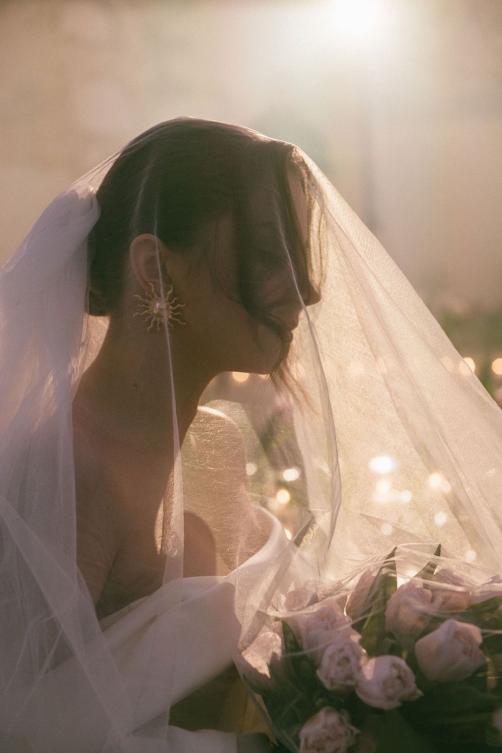 Bride in veil holding pink roses bouquet, bathed in golden backlight, wearing star earring.