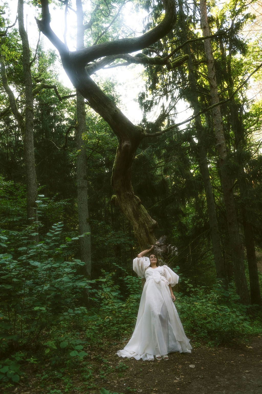 Woman in flowing white gown poses gracefully in a lush green forest beneath dramatic twisted tree branches.