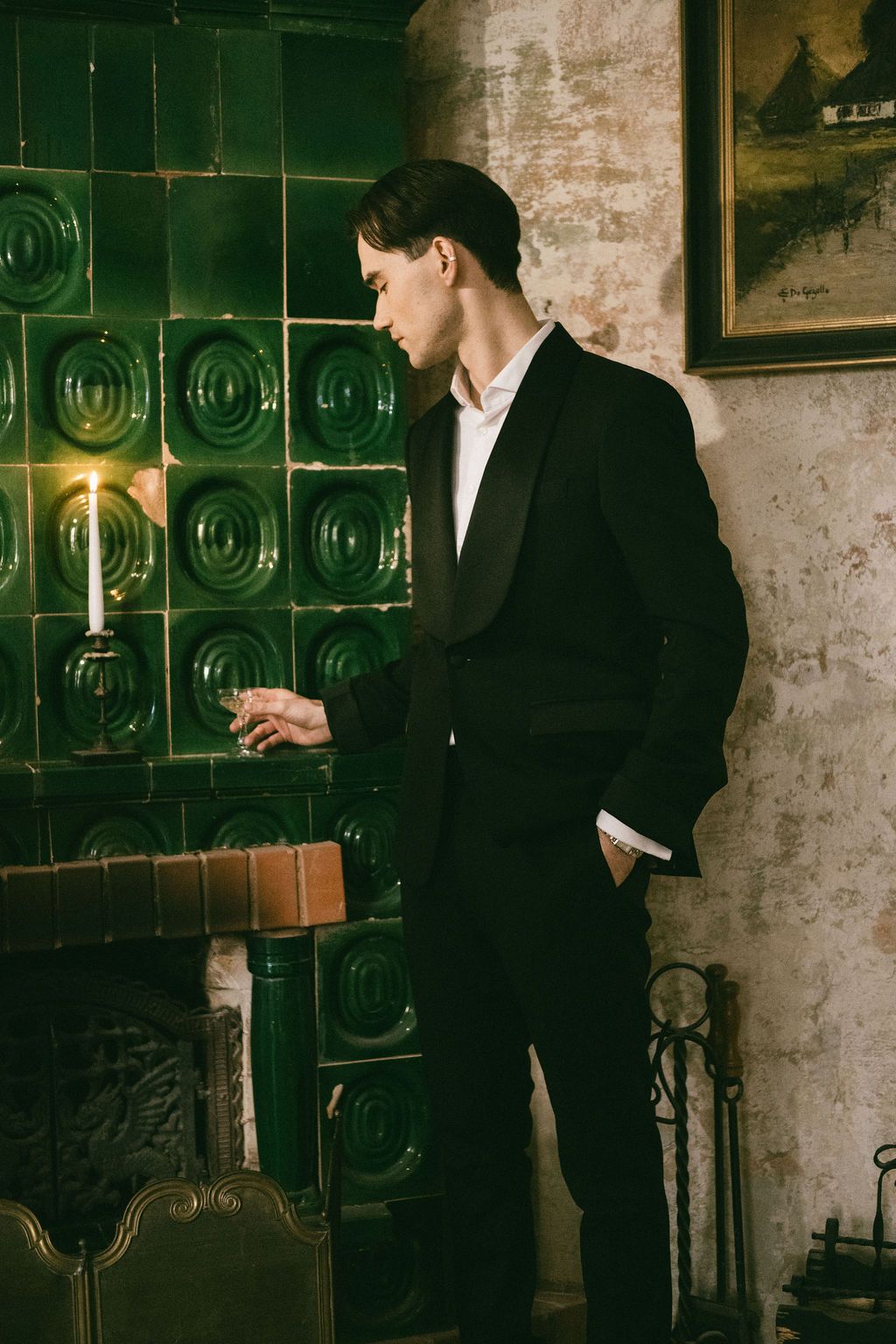 Elegant man in black tuxedo standing by green tiled fireplace with candle in vintage room.