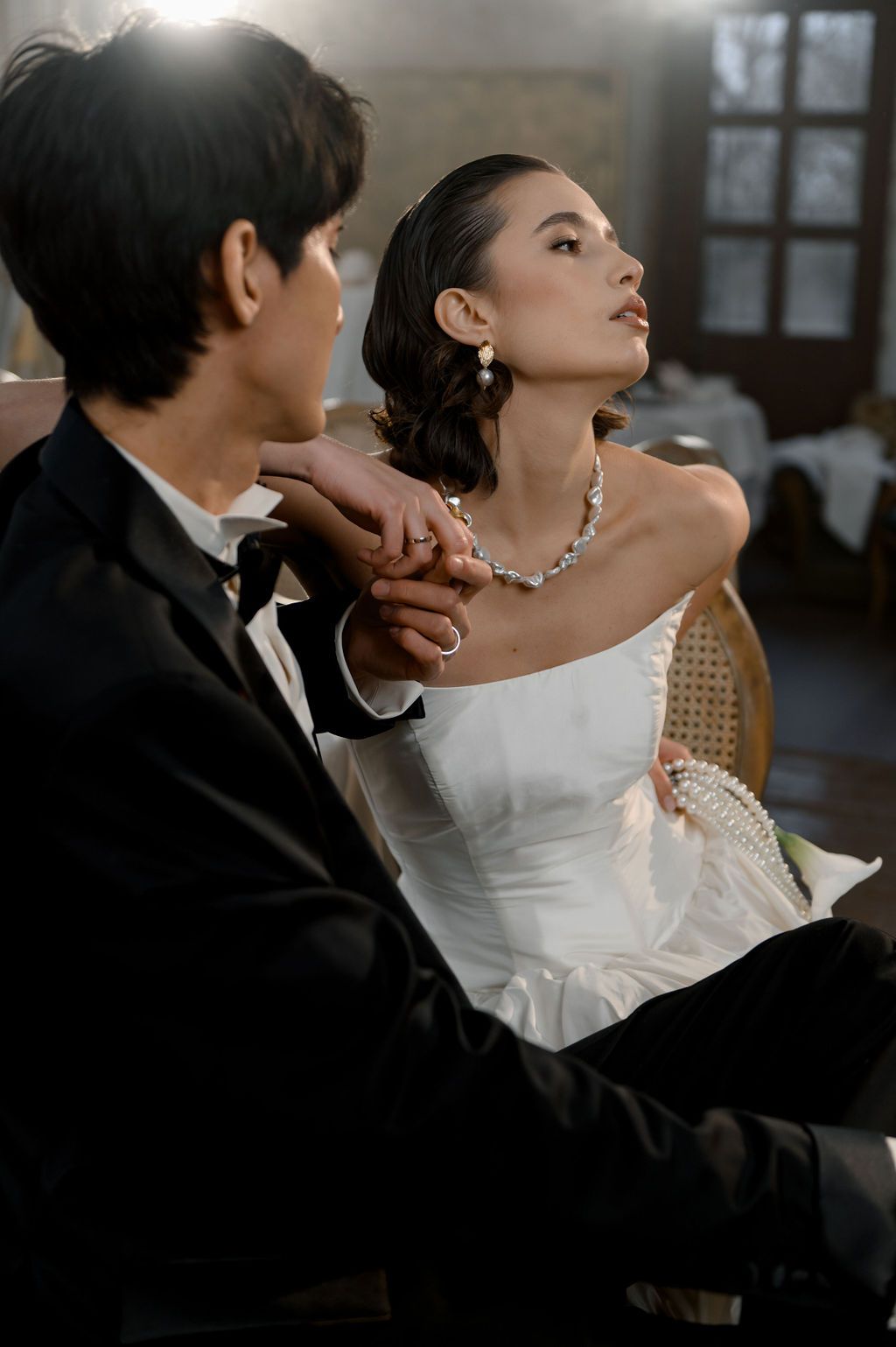 Bride in strapless white gown with pearl necklace holds groom's hand in black tuxedo during elegant wedding ceremony.