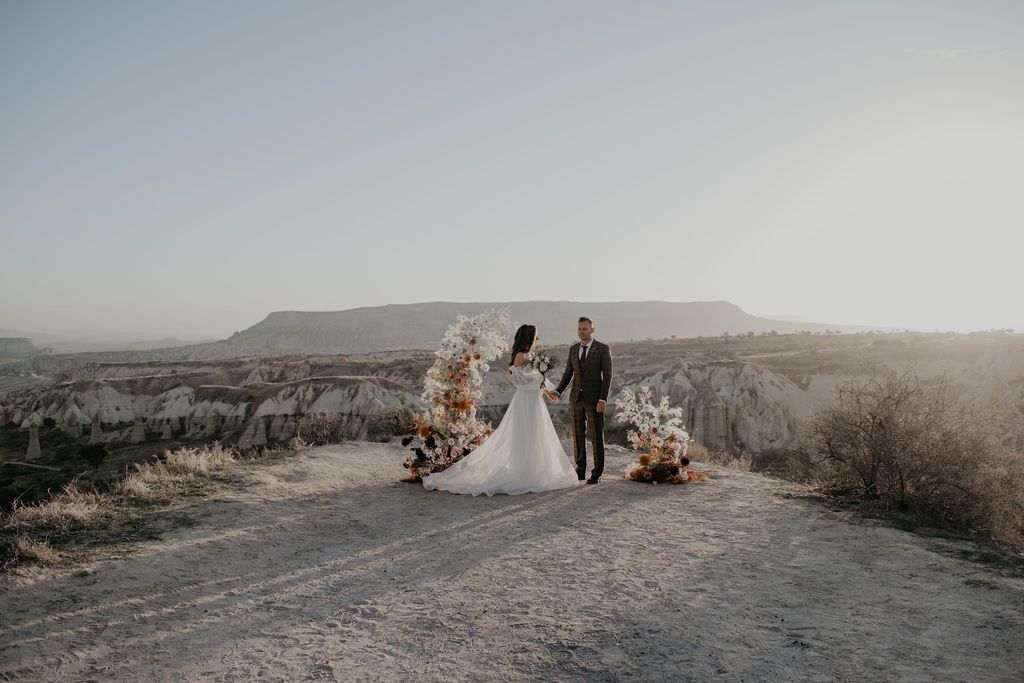Bride and groom standing at floral arch on desert road with scenic rocky landscape backdrop.