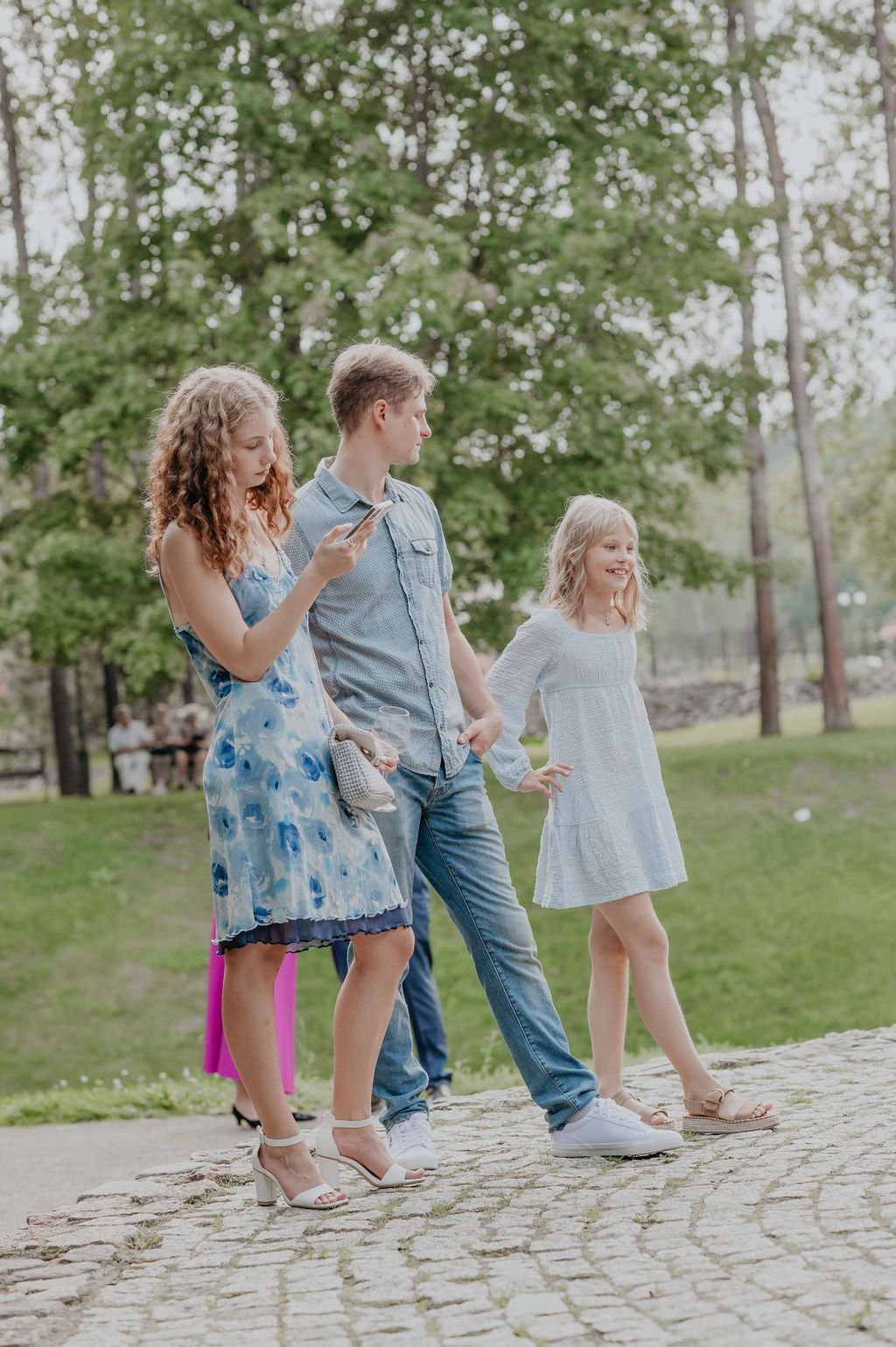 A family of three walks on a cobblestone path in a lush green park, dressed in casual summer outfits.