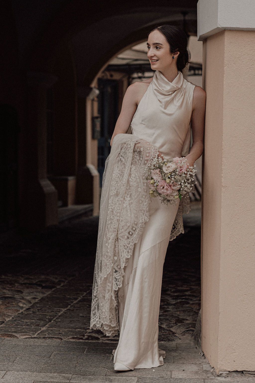 Elegant bride in a sleeveless satin halter gown holds a pink bouquet while leaning against a column in an archway.