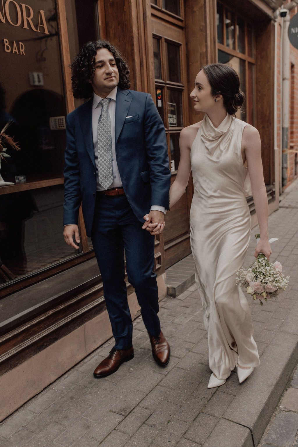 Elegant couple walks hand in hand on cobblestone street, bride in satin gown holding a bouquet.