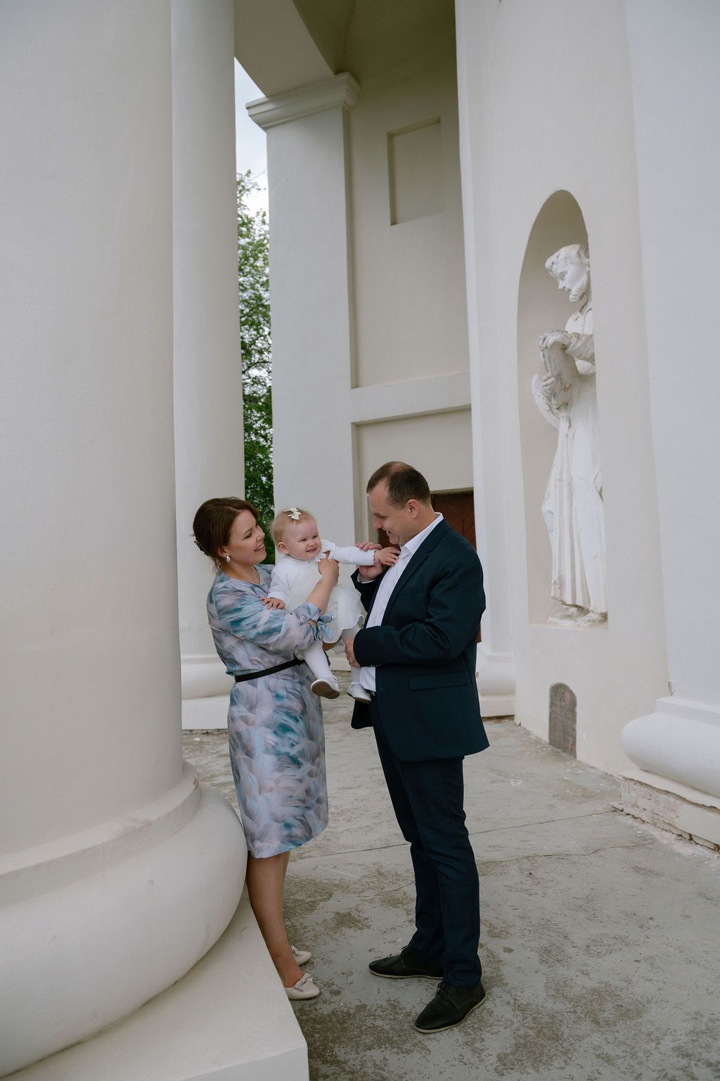 A couple in formal attire holds a baby outside a white neoclassical building with columns and a statue.