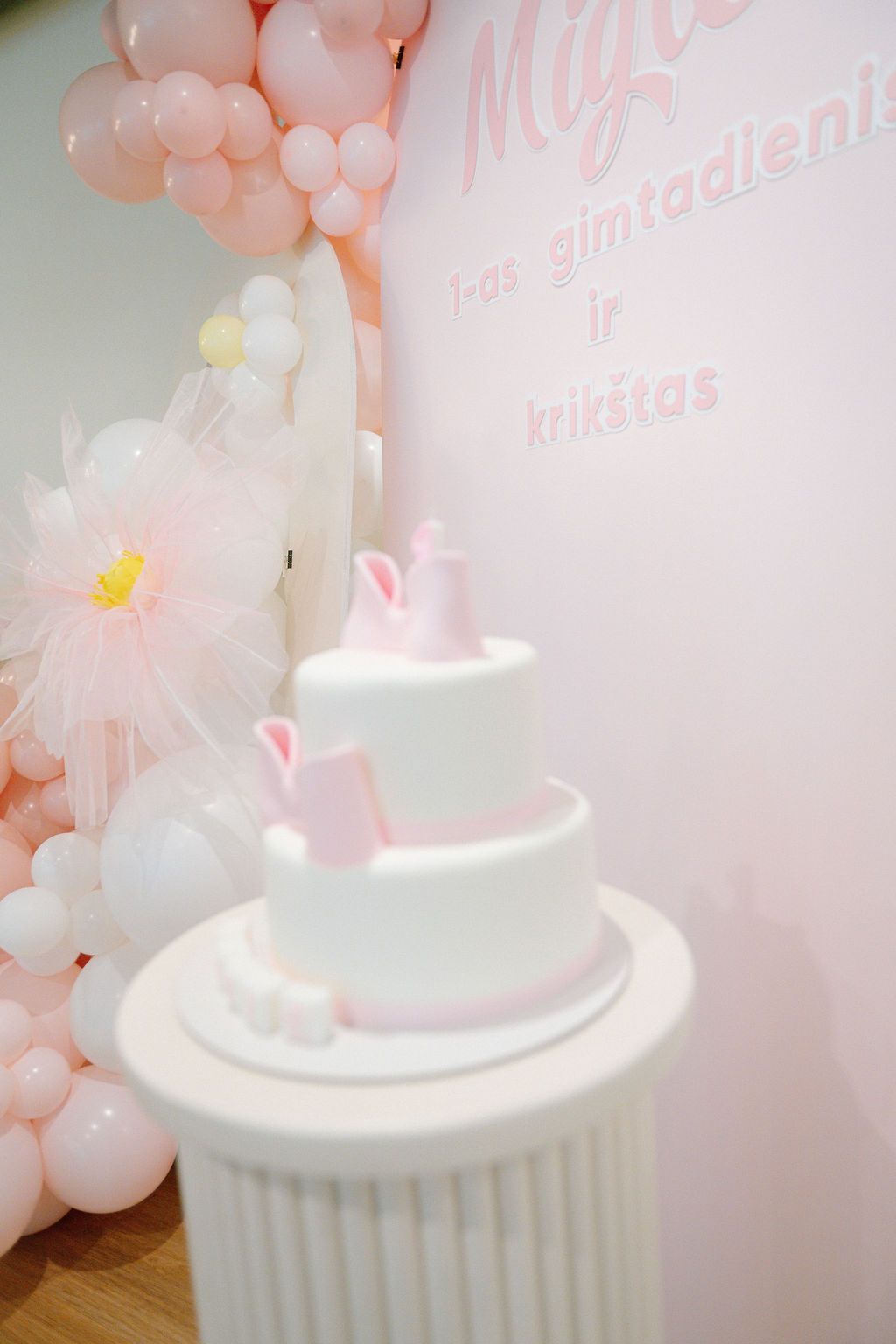 Elegant white three-tier baby birthday cake with pink decorations, displayed on a pedestal surrounded by pink balloon arch.